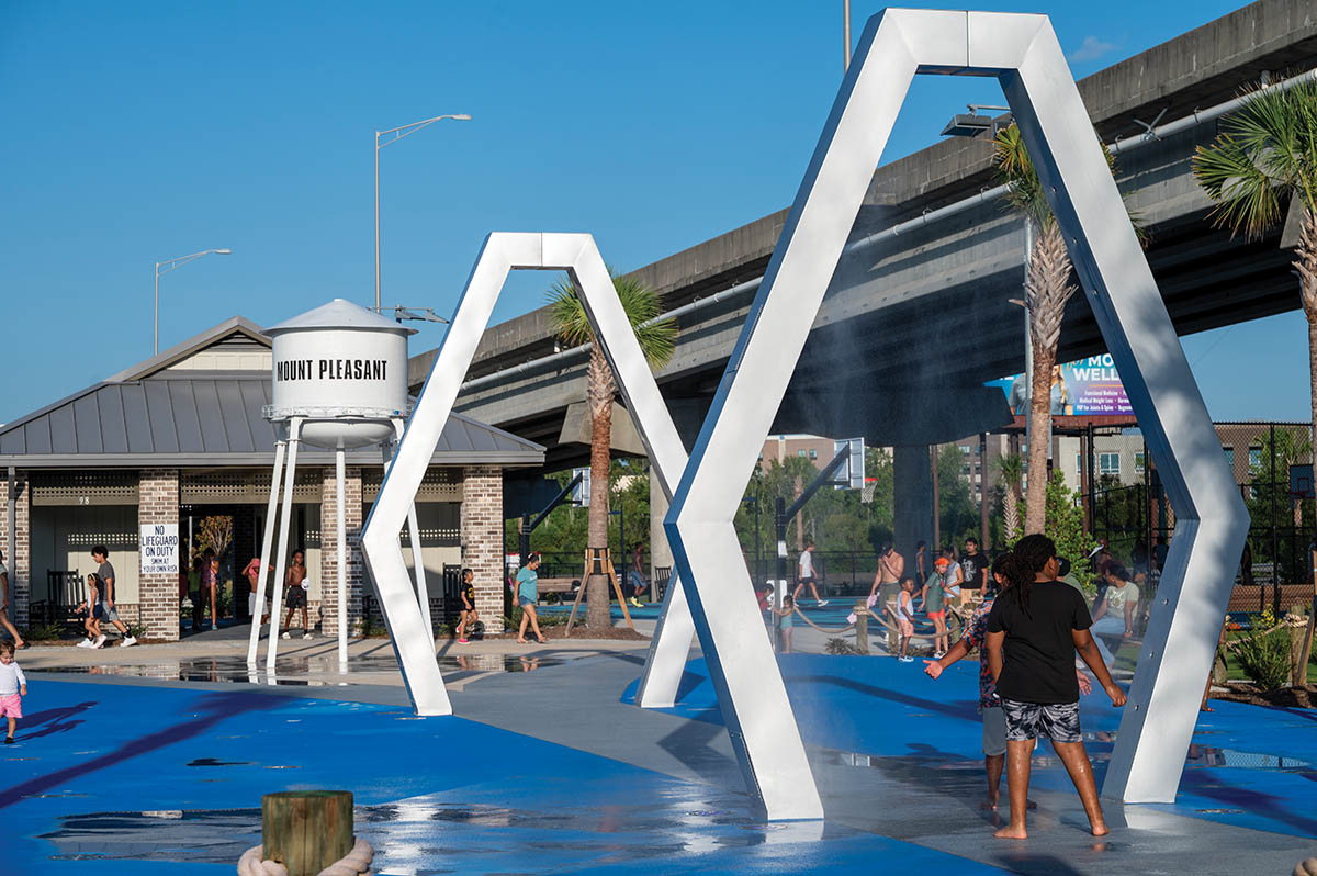 A restroom building with rocking chairs looks out across the splash pad, with sports courts and a fitness area  close by.